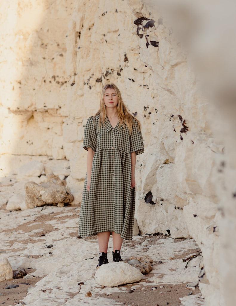 Woman in a green checkered dress standing on a rocky beach with a stone wall in the background